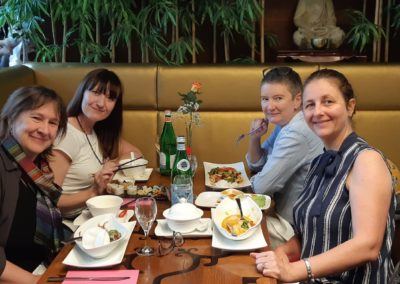 Four women smiling around a wooden restaurant table sharing a meal, with bamboo plants and a Buddha statue behind