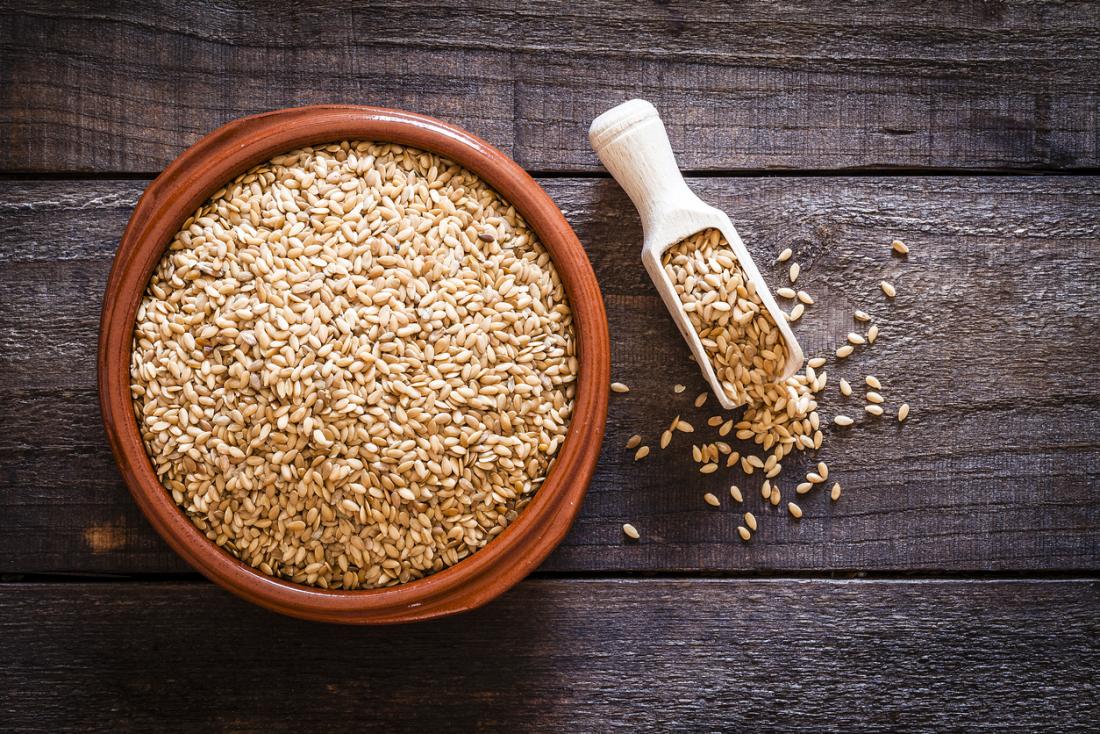 Golden flaxseeds in a terracotta bowl with a wooden scoop spilling seeds onto a rustic wooden table