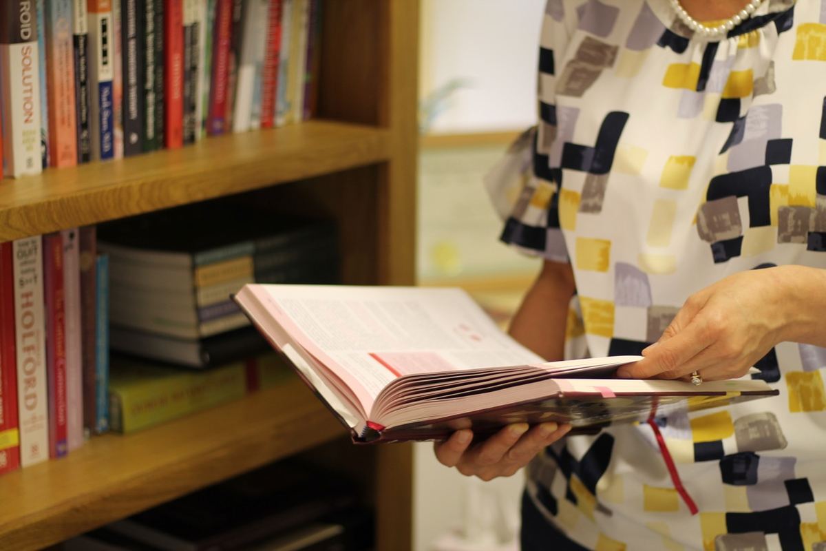 Woman in a patterned blouse and pearl necklace reading an open book beside a wooden bookshelf