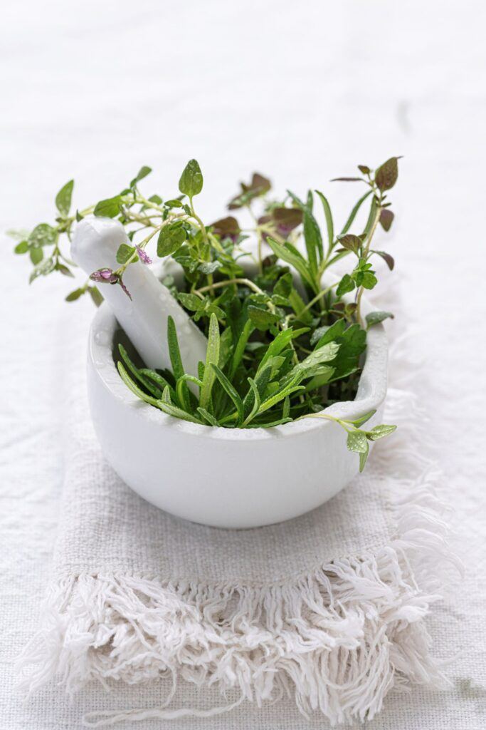 White mortar and pestle filled with fresh green herbs, including rosemary and thyme, on a fringed linen cloth