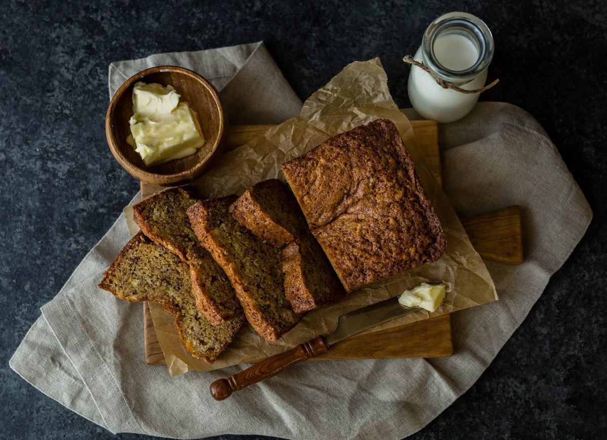 Sliced banana bread loaf on a wooden board with butter, a knife and a bottle of milk