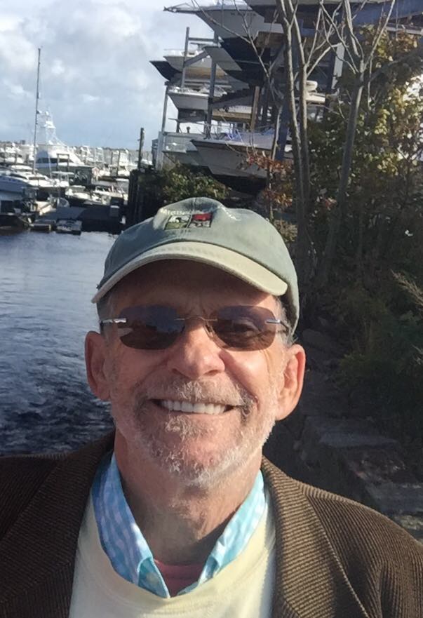 Smiling older man in green cap, tinted glasses and brown blazer, with marina and moored boats behind him