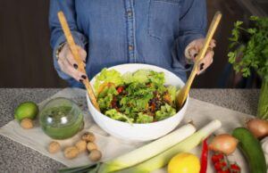 Person tossing a fresh green salad in a white bowl, surrounded by leeks, lemon and vegetables