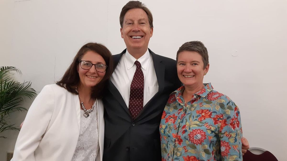 Smiling man in dark suit and burgundy tie posing between two smiling women in a bright room