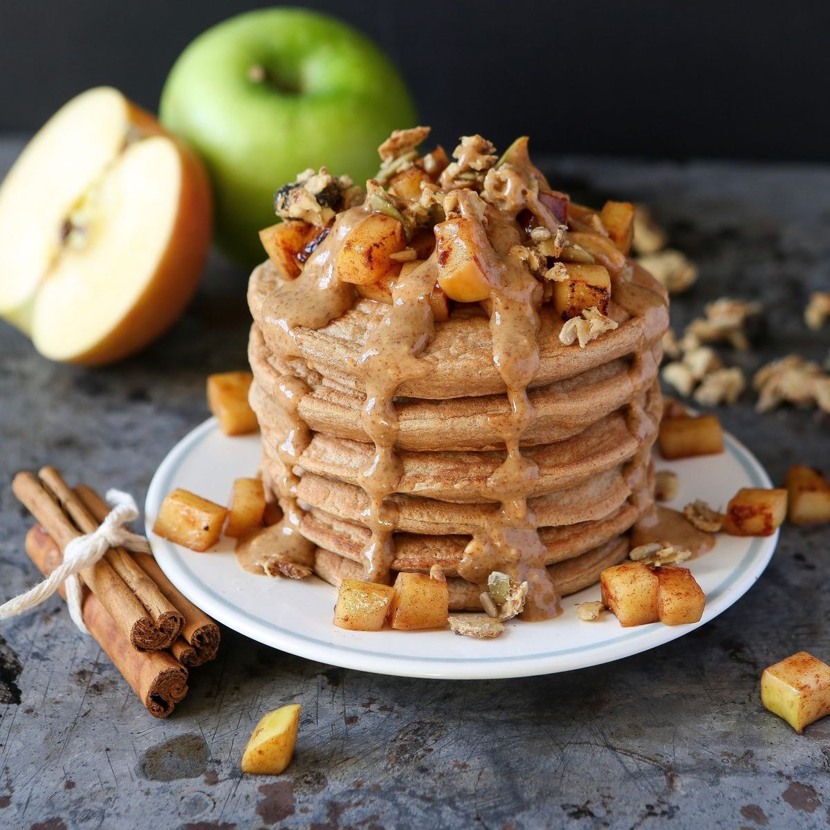Stack of pancakes topped with almond butter drizzle, cinnamon-spiced diced apple and granola, beside cinnamon sticks and apples