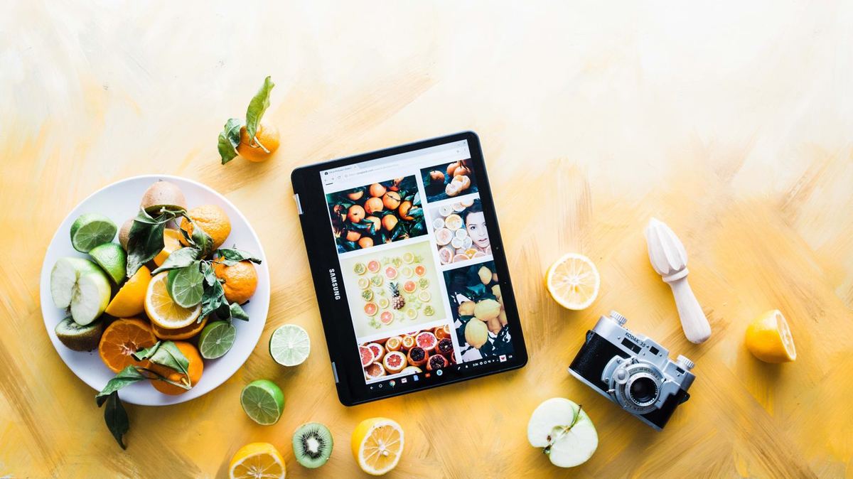 Samsung tablet showing citrus fruit photos, surrounded by halved lemons, limes, kiwi, apples, a vintage Kodak camera and wooden juicer on yellow background