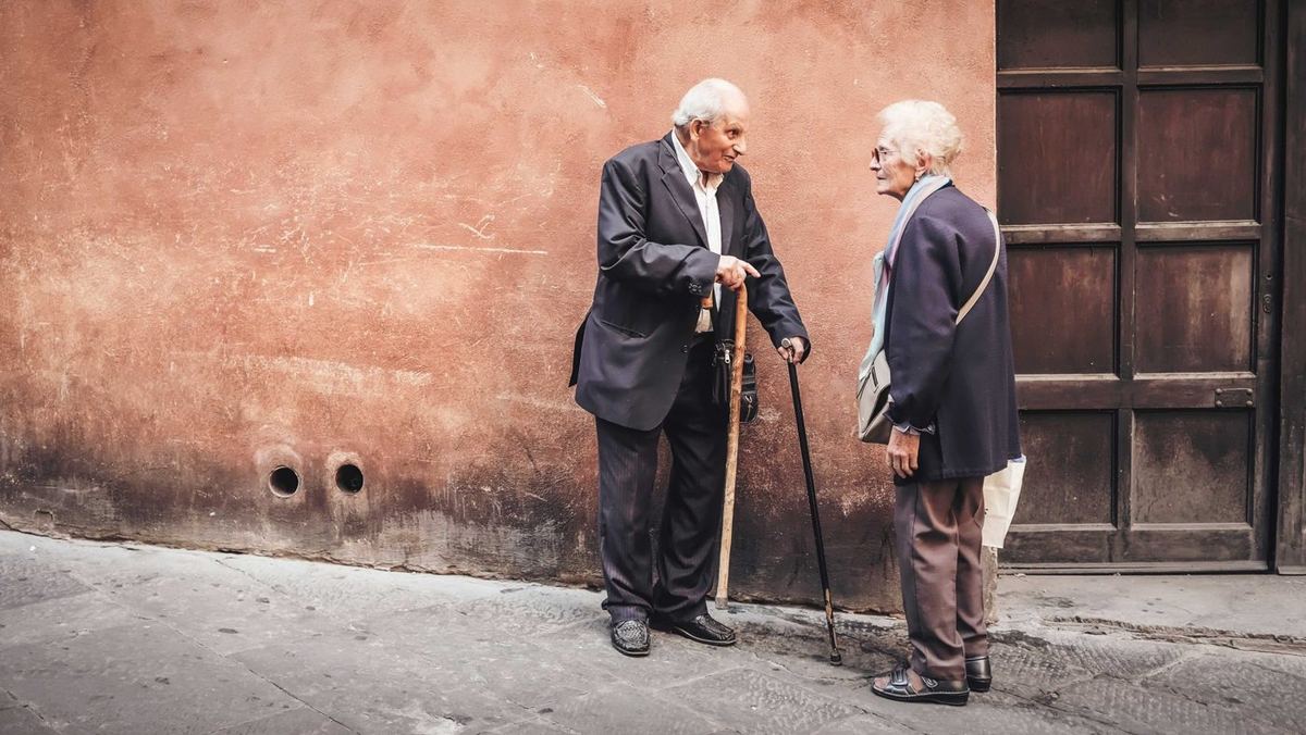 Elderly man and woman talking on a quiet street beside a terracotta wall, he holding two walking sticks