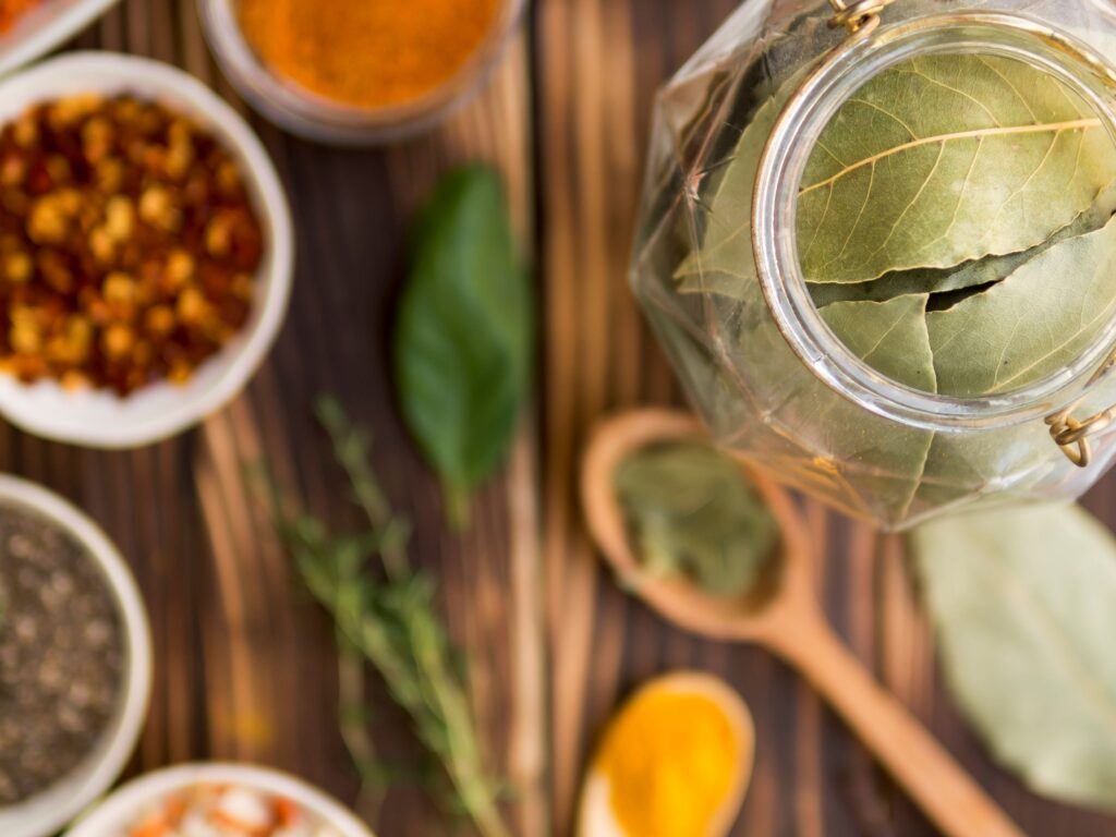 Dried bay leaves in a glass jar surrounded by bowls of spices, thyme and wooden spoons on a rustic table