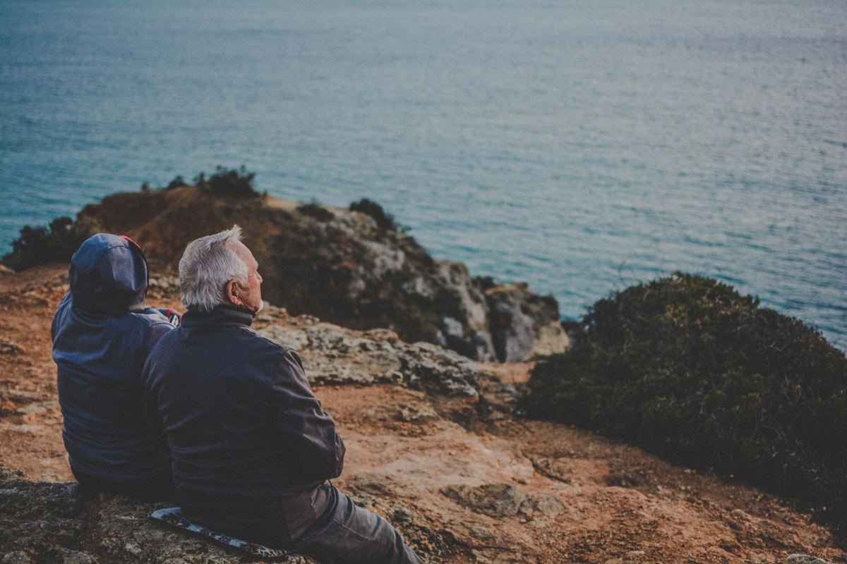 Two people in dark jackets sit close together on a rocky clifftop, gazing out at the calm sea