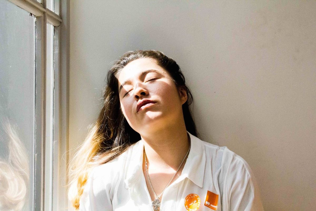Woman with eyes closed and head tilted back against a wall, resting in a patch of sunlight beside a window