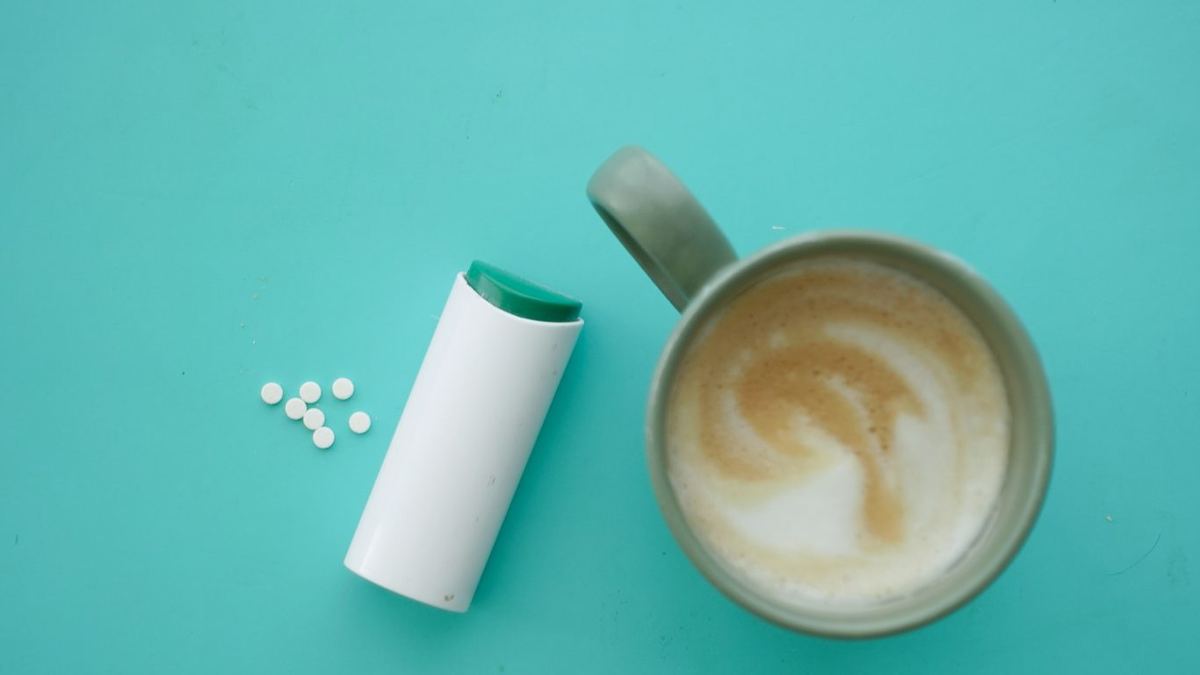 Sweetener tablets spilled from a white dispenser beside a mug of milky coffee on a turquoise surface