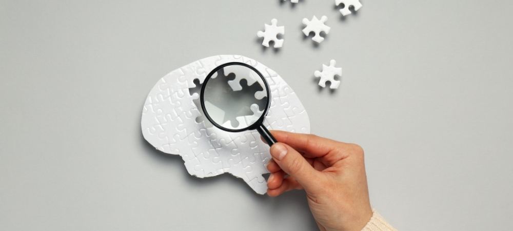 Hand holding magnifying glass over white jigsaw puzzle shaped like a brain with missing pieces