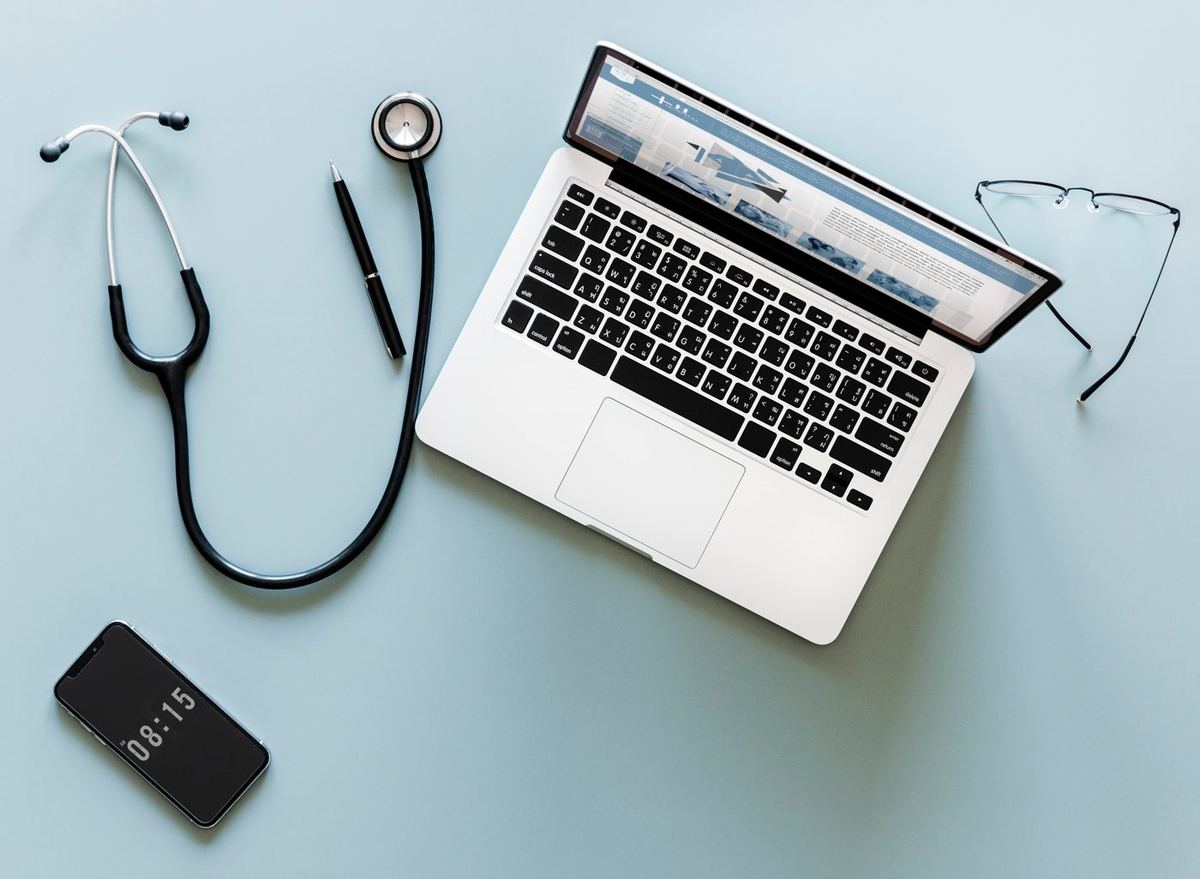 Stethoscope, laptop, pen, glasses and smartphone showing 08:15 arranged on a pale blue desk surface