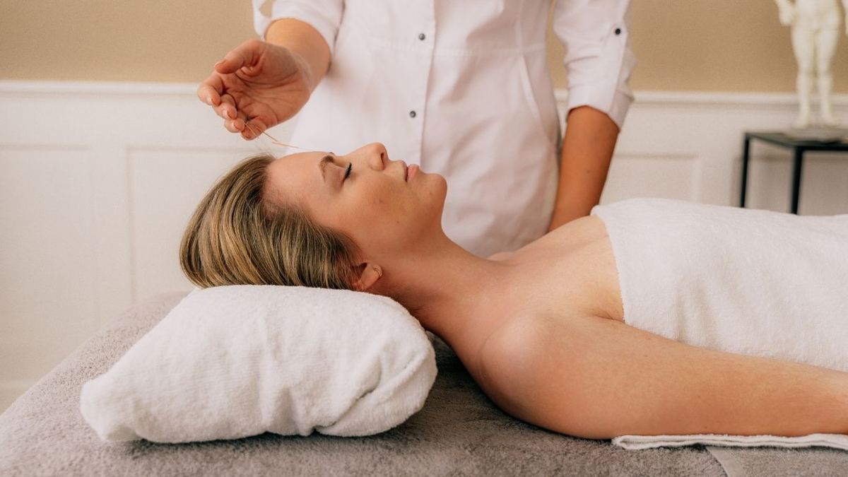 Practitioner in white tunic inserting an acupuncture needle into the forehead of a woman lying relaxed on a treatment couch