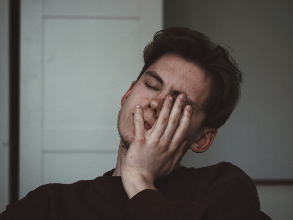 Tired young man with eyes closed, resting his face heavily in one hand, looking mentally exhausted
