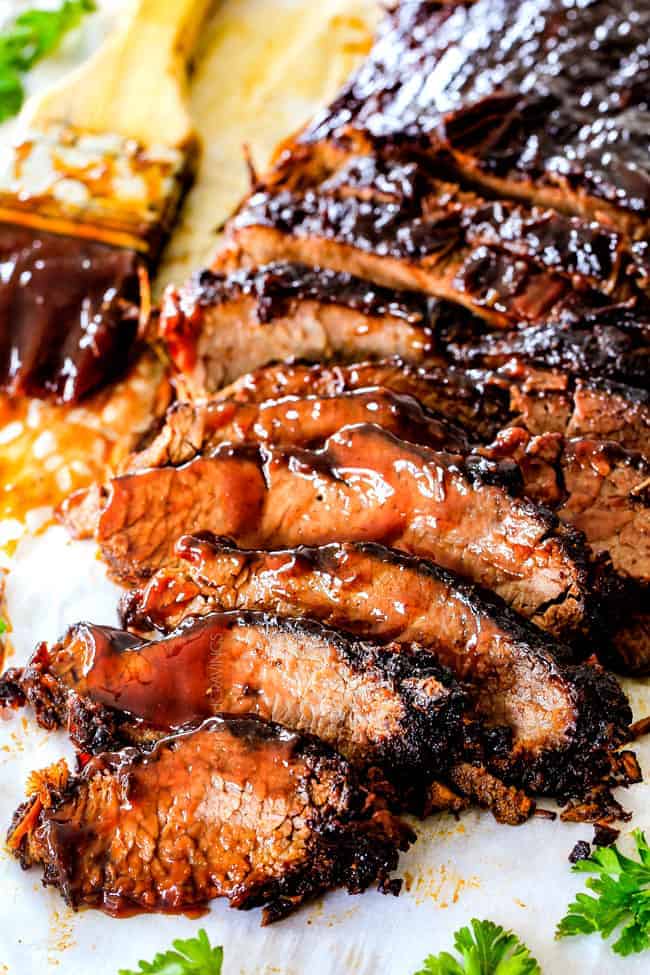 Sliced barbecue-glazed beef brisket on baking paper with a basting brush and fresh parsley