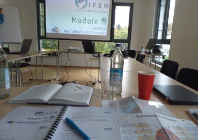 Training room set up for an IFEN neurofeedback course, with open workbook, water bottle and projected module slide